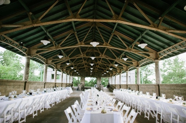 Outdoor wedding reception venue with long tables and white chairs under a green and wood pavilion.
